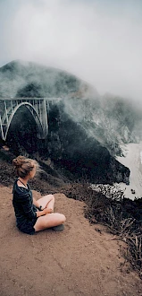 A person sits on a cliff overlooking a foggy coastline with a bridge in the background, capturing a serene and scenic moment.