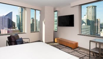 A modern hotel room with a large bed, flat-screen TV, armchair, and a view of city skyscrapers through multiple windows.
