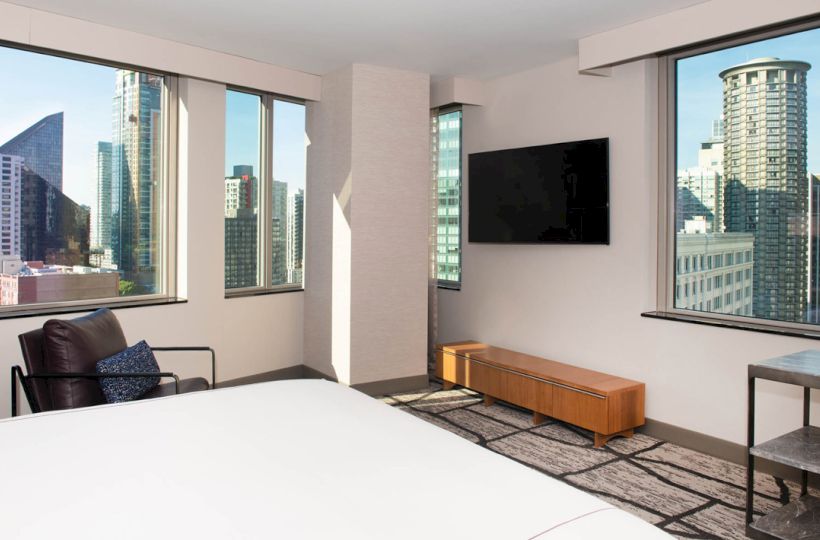 A modern hotel room with a large bed, flat-screen TV, armchair, and a view of city skyscrapers through multiple windows.