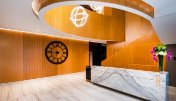 A modern lobby with a marble reception desk, large wall clock, wooden paneling, and elegant lighting fixture above.