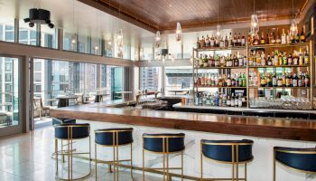 A modern bar with a wooden ceiling, featuring a long counter, stylish bar stools, and a wall of liquor bottles against large windows.