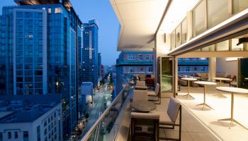 A modern balcony overlooking a city street, with tables and chairs, adjacent to tall buildings at dusk, under a blue sky.