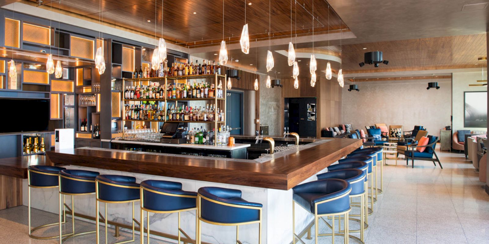 A modern bar with a central counter, blue bar stools, hanging lights, and shelves stocked with bottles. Comfortable seating in the background.