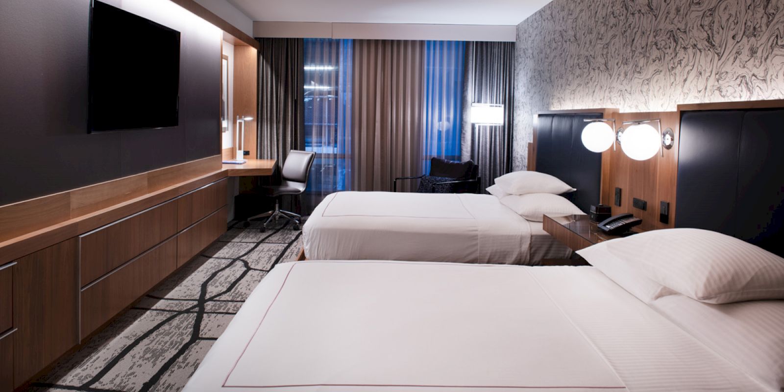 The image shows a hotel room with two beds, a wall-mounted TV, desk, chair, and modern decor.