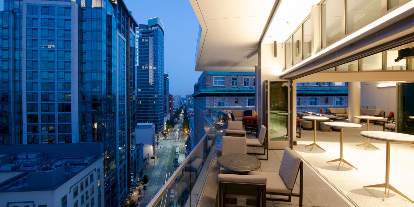 A modern, lit balcony with tables and chairs overlooks a city street lined with tall buildings at dusk.