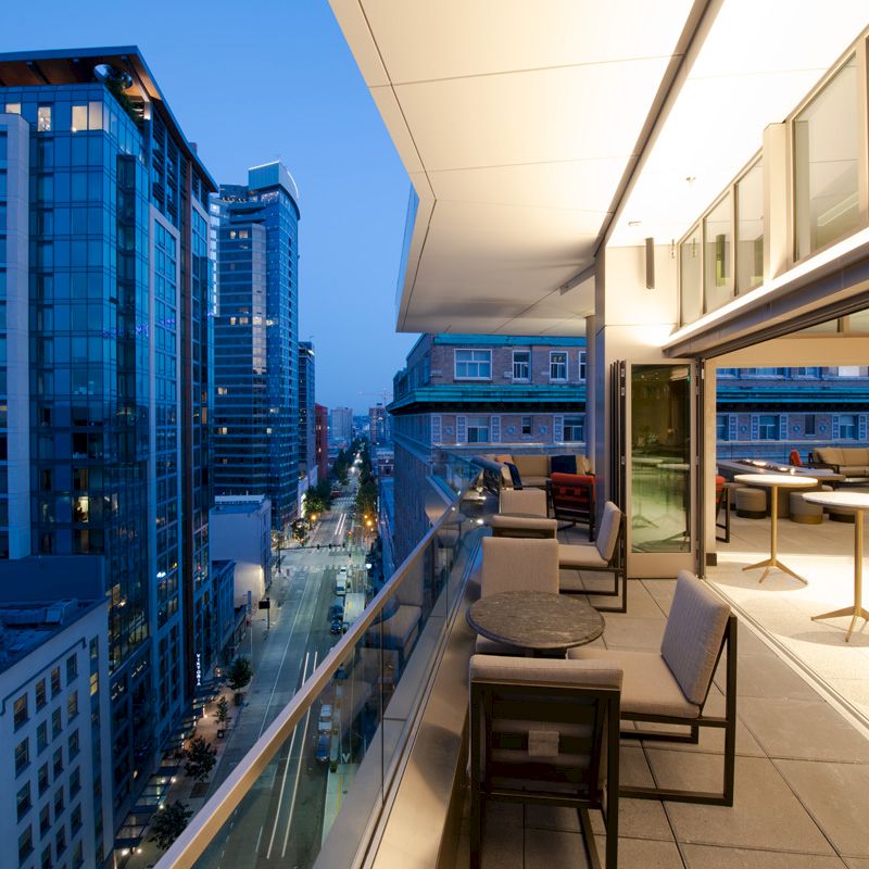 A modern cityscape at dusk is seen from a terrace with tables and chairs, overlooking tall, illuminated buildings and a street below.