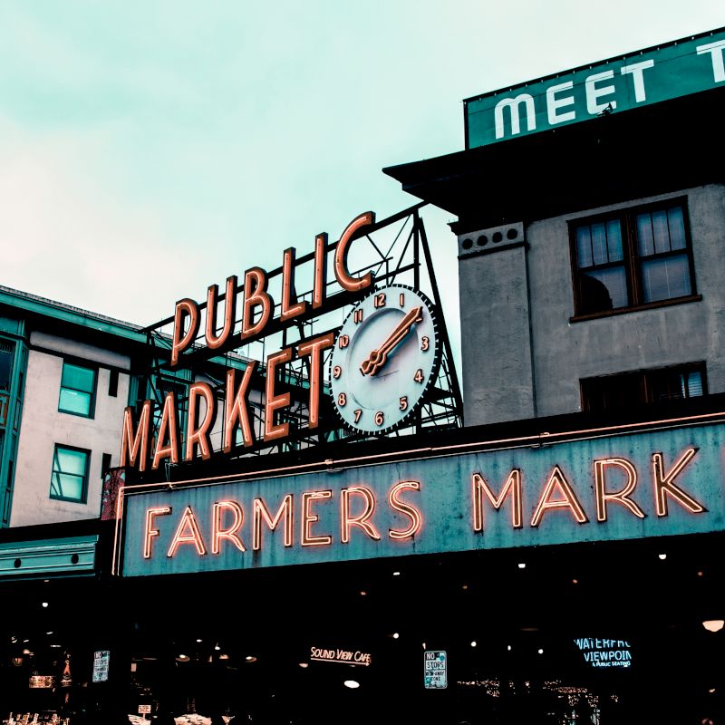 The image shows a public market sign with a clock and neon lights reading "FARMERS MARKET" on a building under a cloudy sky.