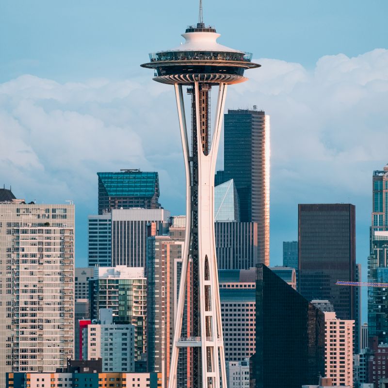 The image shows the Space Needle with a backdrop of Seattle's cityscape, featuring numerous tall buildings under a cloudy sky.