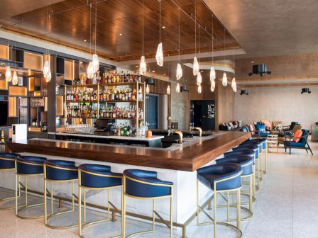 A modern bar with blue chairs, hanging lights, and a stocked back wall. Contemporary design with seating area visible in the background.