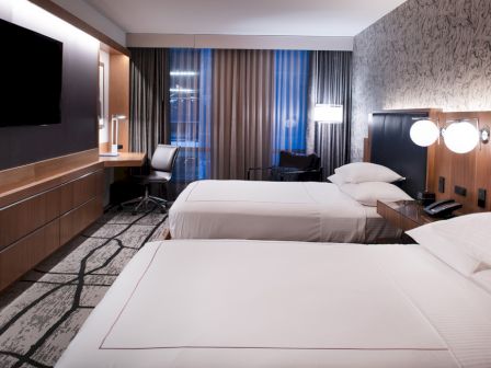 The image shows a modern hotel room with two beds, a wall-mounted TV, a desk with a chair, and stylish lighting and decor.