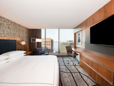 A modern hotel room features a large bed, a flat-screen TV, a desk, and a view of city buildings through a big window.