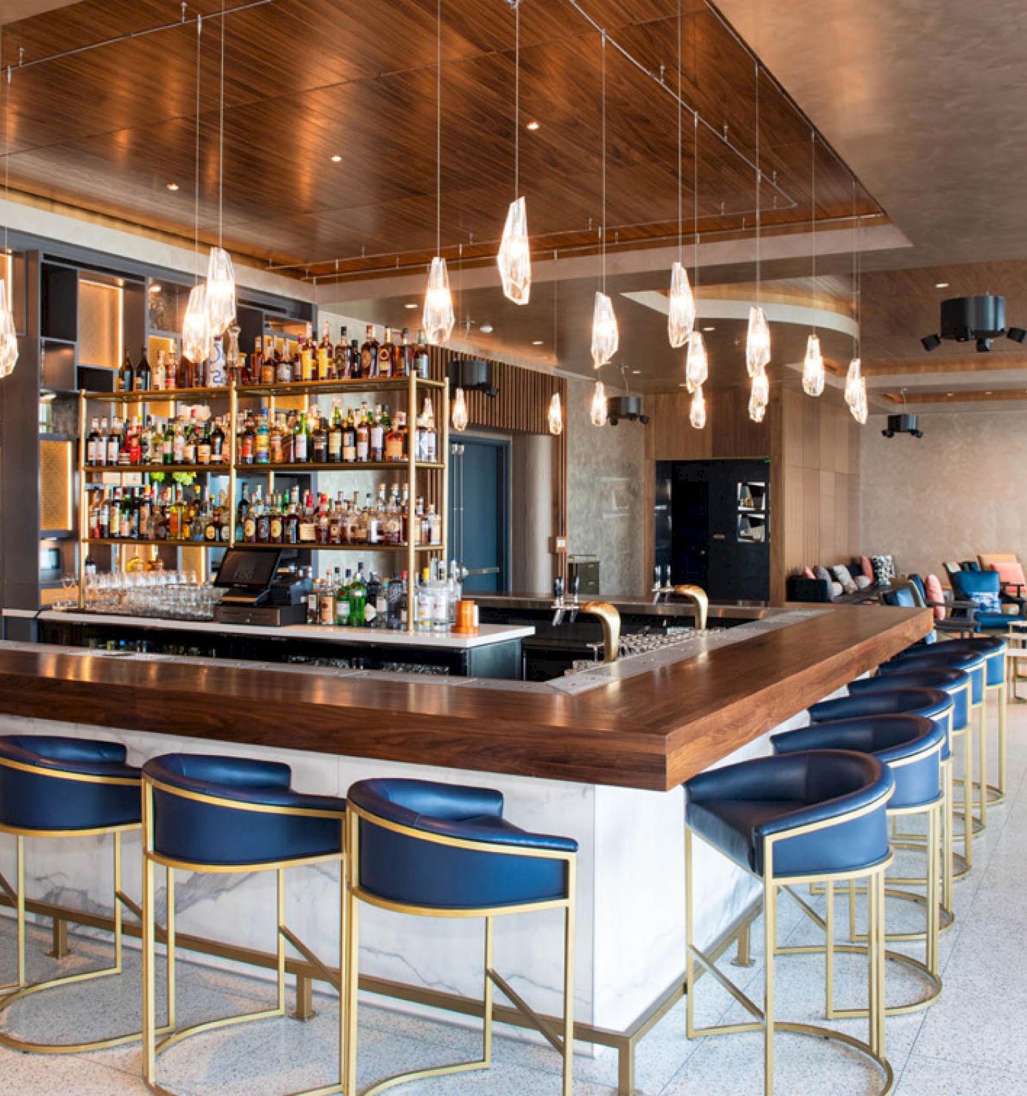 A stylish bar with a central counter, blue stools, hanging lights, and a wide selection of bottles. Comfortable seating is seen in the background.