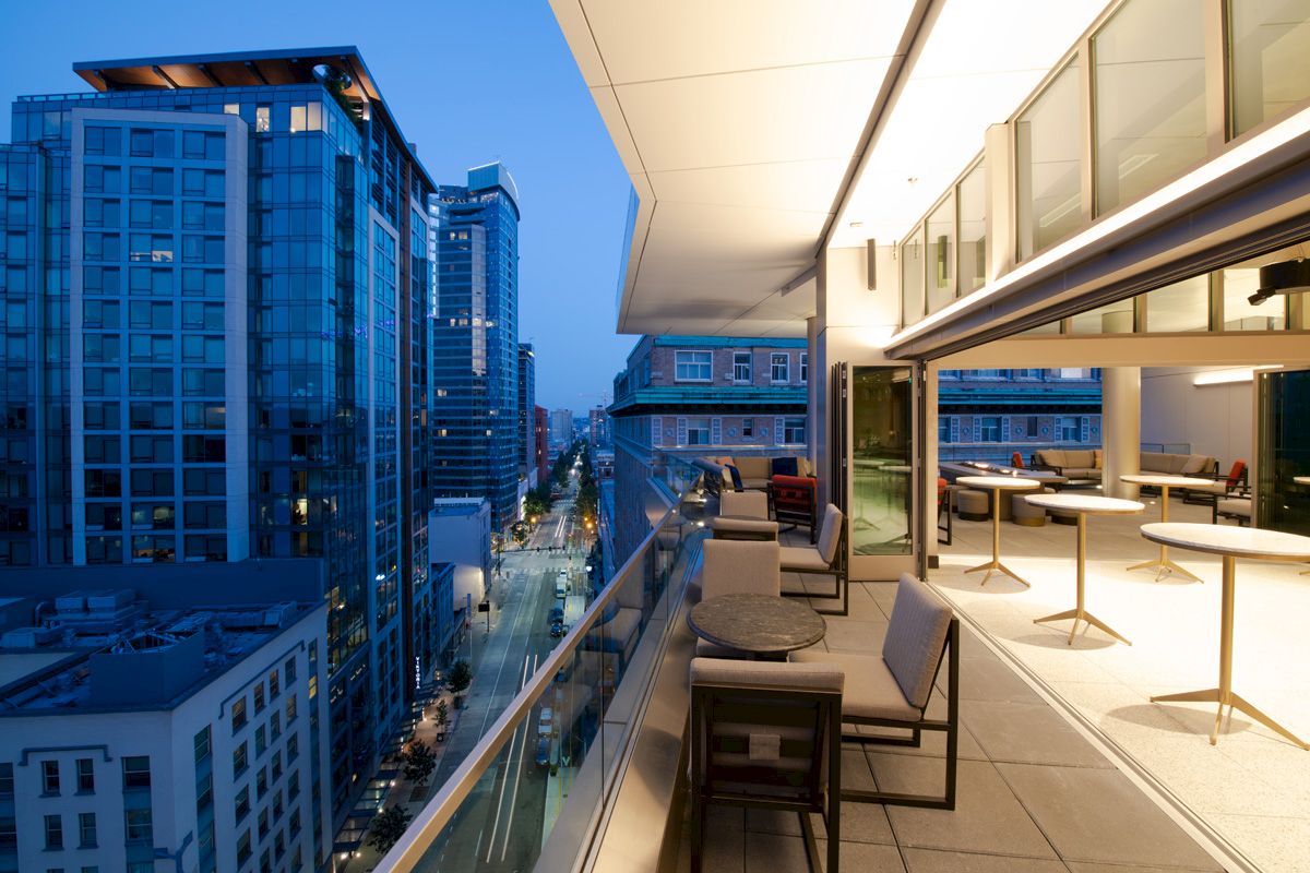 A modern cityscape at dusk with tall buildings and a stylish balcony with seating, overlooking a street.
