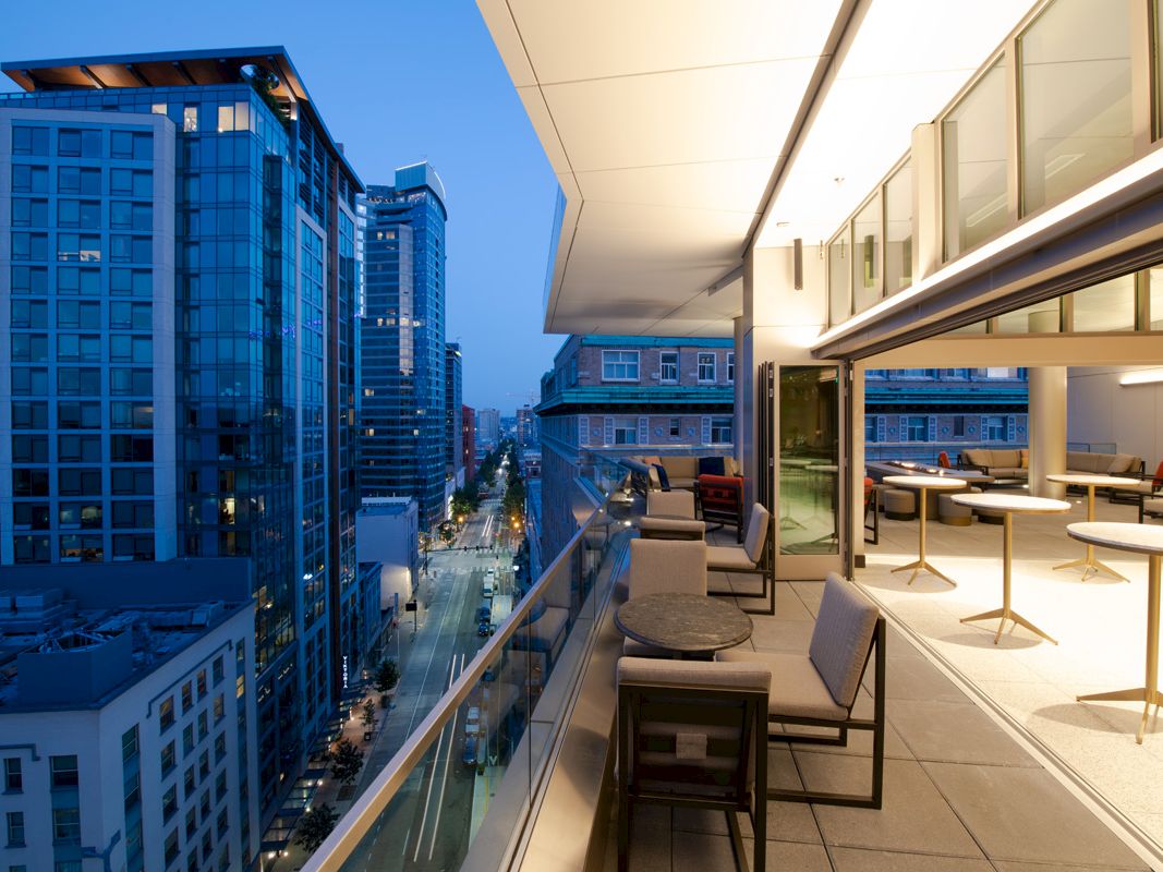 A modern cityscape at dusk with tall buildings and a stylish balcony with seating, overlooking a street.