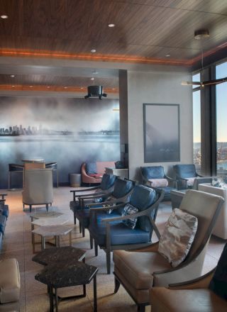 The image shows a modern lounge with sleek seating, large windows offering waterfront views, and a mural of a city skyline on the wall.