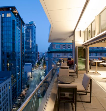The image shows a modern urban balcony at dusk with chairs and tables, overlooking a city street with tall buildings and illuminated windows.
