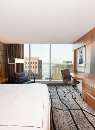 The image shows a modern hotel room with a bed, a large TV, a desk, and a city view through a window, featuring contemporary decor.
