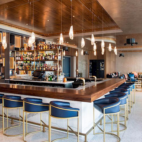 The image shows a modern bar with blue stools, hanging lights, a stocked bar, and seating area in the background.