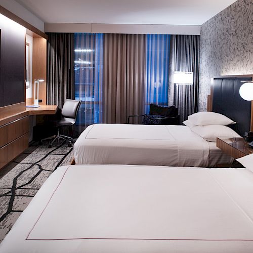 The image shows a modern hotel room with two beds, a wall-mounted TV, a desk and chair, and stylish lighting. Decor is contemporary.