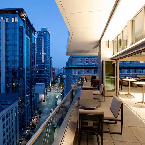 A modern rooftop terrace at dusk with seating and tables, overlooking a city street with tall buildings and illuminated windows.