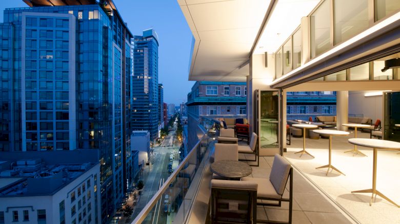 A rooftop terrace overlooking a city street with modern buildings at twilight, featuring outdoor tables and chairs.