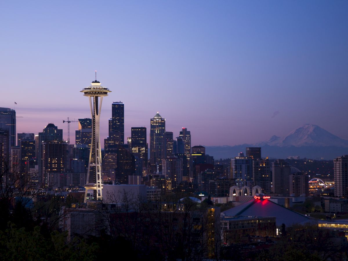 The image shows a city skyline at dusk with the Space Needle and Mount Rainier in the background, likely Seattle, under a colorful evening sky.