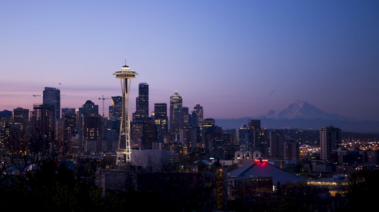 The image shows a city skyline at dusk with the Space Needle and Mount Rainier in the background, likely Seattle, under a colorful evening sky.