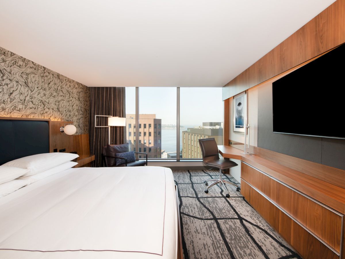 A modern hotel room with a large bed, flat-screen TV, work desk, and large window overlooking a cityscape with buildings.