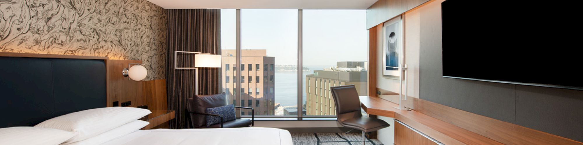 The image shows a modern hotel room with a large bed, a desk, a TV, and a view of buildings and the sea through the window.